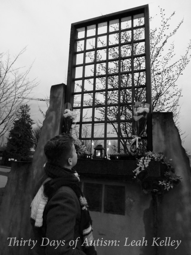 Black and White photo of H looking up at and art installation represent cement walls with the barred windows of Woodlands Institution. This is has been recreated so that it is the same height that the windows would have been for the residents. There are candles and flowers arranged on the window's ledge, and a tree can be seen through the bars.
