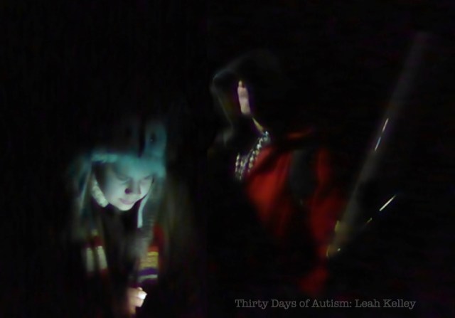 "Cosplay Stargazers" Image: H looking downwards, dressed in vintage wool trenchcoat, Dr Who scarf and owl hat and K standing behind, mostly hidden by darkness and a hooded cape over a red/orange jacket. K appears to be holding a wooden staff. The background is night black with lighting from below. 